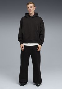 Ung mand står mod en grå baggrund iført sort Puma-hættetrøje, sorte bukser med vide ben og sorte Puma-sneakers.