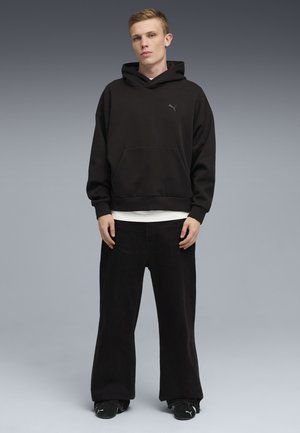 Giovane uomo in piedi contro uno sfondo grigio, indossa una felpa nera Puma con cappuccio, pantaloni neri a gamba larga e sneakers nere Puma.