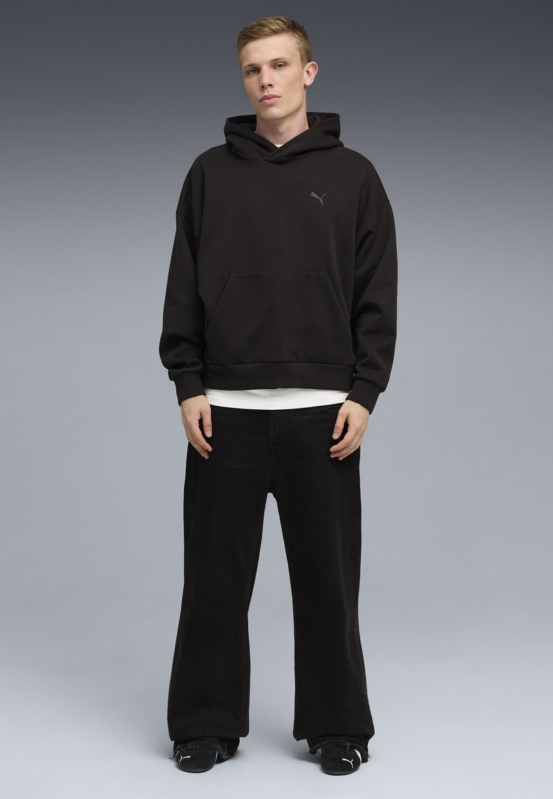 Ung mand står mod en grå baggrund iført sort Puma-hættetrøje, sorte bukser med vide ben og sorte Puma-sneakers.