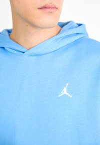 Gros plan sur une personne portant un sweat à capuche bleu clair avec un petit logo silhouette blanc d'un joueur de basket en plein saut sur la poitrine.