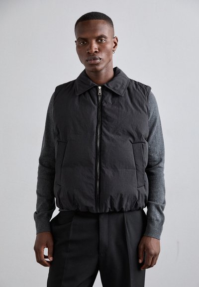 Črn puffer brezrokavnik z zadrgo spredaj, velikimi stranskimi žepi in ovratnikom. Obutel je preko sivega dolgih rokavov, teksturirane tkanine s prilagojeno zasnovo.