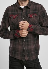 Chemise à carreaux marron avec des accents rouges, deux poches poitrine et un col boutonné. Fabriquée en tissu doux et texturé.
