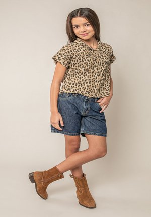 Meisje staat met een been gekruist, draagt een kortärmelig overhemd met luipaardprint, spijkershort en bruine suède enkellaarsjes.