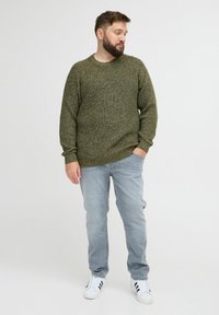 Olivenfarbener Strickpullover mit Rundhalsausschnitt, kombiniert mit hellblauen Jeans und weißen Sneakers. Lockere Passform mit gerippten Ärmelbündchen.