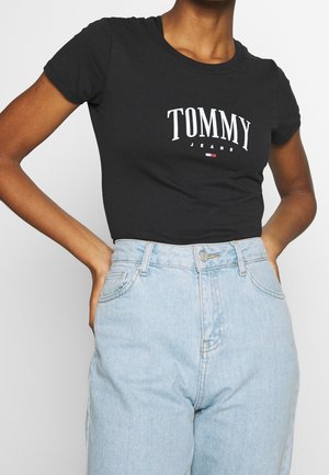 Personne portant un t-shirt noir ajusté à manches courtes de Tommy Jeans rentré dans un jean bleu clair taille haute, les mains sur les hanches.