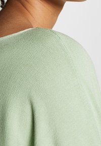 Suéter de punto verde claro con una textura suave, que presenta un escote redondeado y un distintivo detalle de punto acanalado a lo largo del borde.