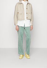 Beige kurze Jacke mit großen Knöpfen, weißes Hemd, strukturierter Pullover, mintgrüne Cordhosen und gelbe Plateauschuhe.