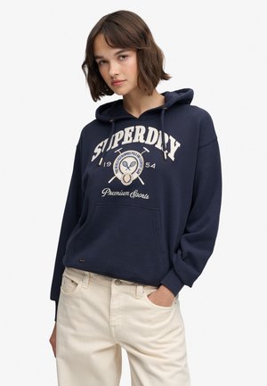 Ung kvinde iført en mørkeblå Superdry hættetrøje med sportslogo og cremefarvede bukser, stående med den ene hånd i lommen mod en hvid baggrund.