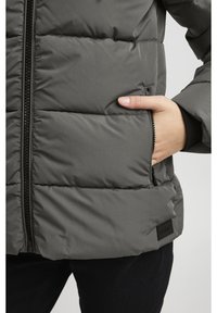 Szara puchowa kurtka na zamek, z poziomymi pikowanymi panelami, boczną kieszenią oraz małą naszywką z logo przy dolnym brzegu.