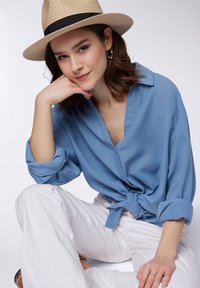 Top azzurro chiaro, morbido e con scollo a V, con dettaglio annodato in vita, abbinato a pantaloni bianchi a gamba larga e un cappello di paglia con una fascia nera.