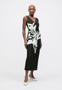 Mulher vestindo um vestido midi preto texturizado sem mangas, com grandes bordados florais brancos, combinado com sapatos de salto branco de bico fino.