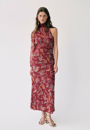 Femme portant une robe rouge à fleurs sans manches avec un col haut et une manche longue en voile transparent, associée à des talons dorés à lanières.