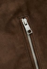 Tela de ante marrón con un primer plano de una cremallera metálica. La cremallera presenta una lengüeta rectangular con un acabado pulido.