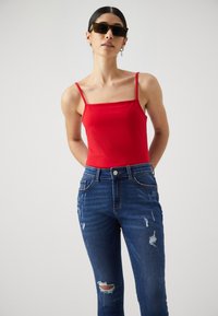 Κόκκινο αμάνικο τοπ με λεπτές τιράντες, συνδυασμένο με τζιν ψηλόμεσα σε μπλε χρώμα με σκισίματα και σκούρα πλύση.