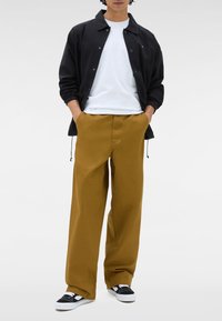 Uomo che indossa una giacca nera sopra una maglietta bianca, pantaloni larghi color senape e sneakers in bianco e nero, con le mani nelle tasche.