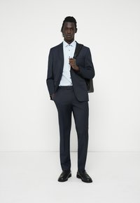 Mörkblå skräddarsydd kostym med fickklaffar, åtföljd av en ljusblå skjorta. Svarta skor och en svart axelväska fulländar outfiten.
