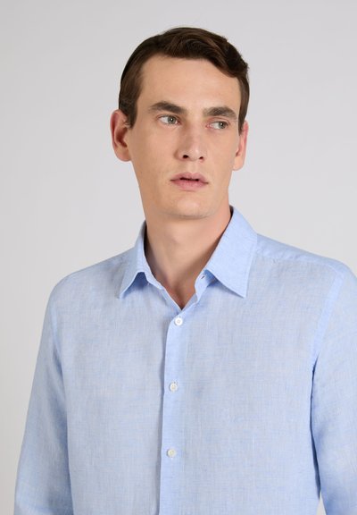 Hombre con una camisa de botones azul claro, mirando ligeramente hacia la derecha, sobre un fondo liso de color gris claro.