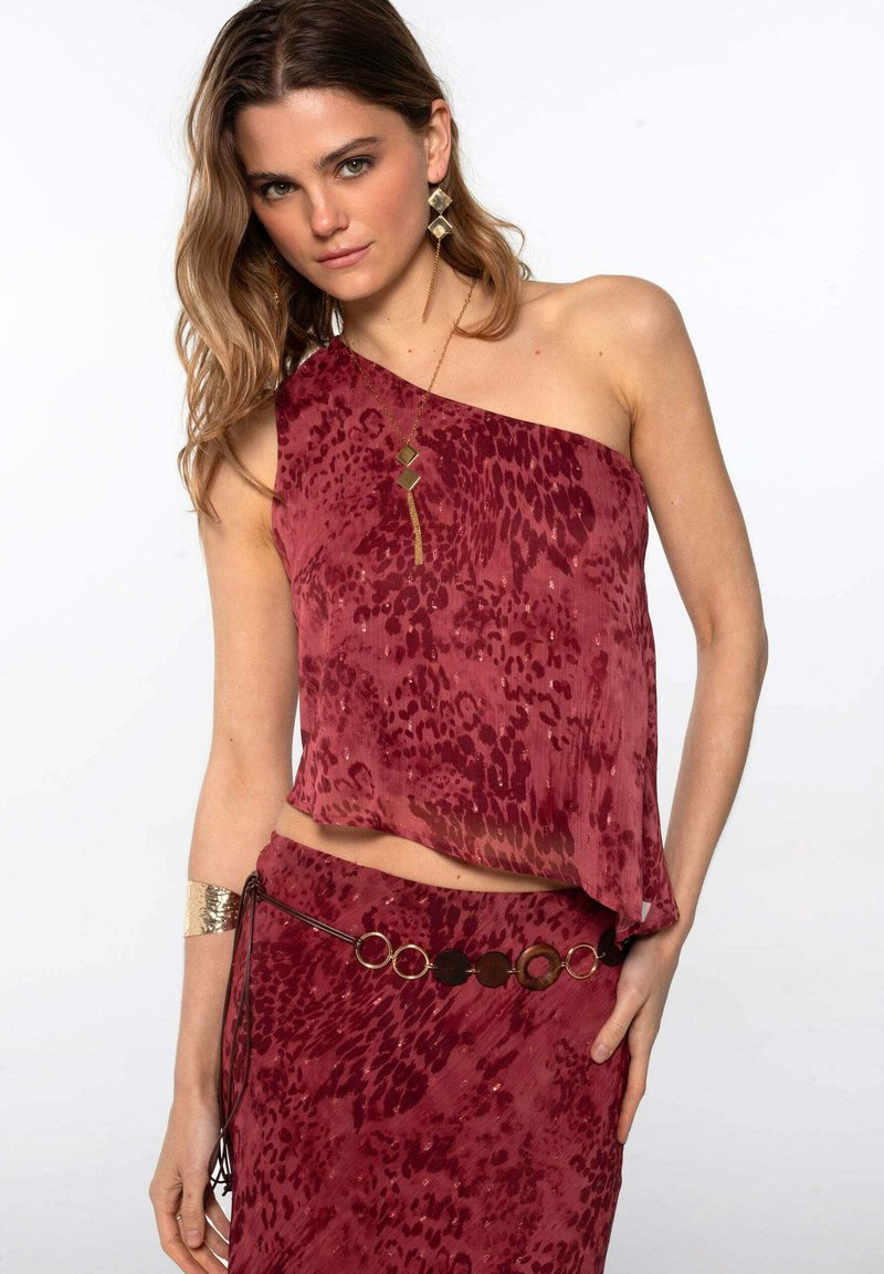 Haut asymétrique bordeaux à motif animalier, assorti à une jupe et une ceinture circulaire accent. Comprend des bijoux dorés et un tissu texturé.