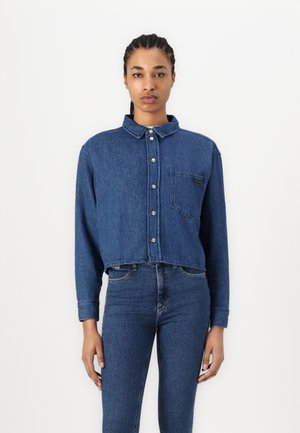 Denim cropped shirt with button closure, collar, and front pocket; deep blue color, slightly faded texture, paired with fitted jeans.