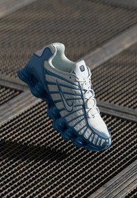 White and blue athletic sneaker with cushioned sole hovering above a textured metal grid surface.