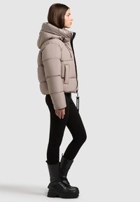 Beżowa kurtka puchowa z dużym kapturem, poziomym pikowaniem, przednią kieszenią i metką z logo, zestawiona z czarnymi legginsami i grubymi czarnymi butami na sznurówki.