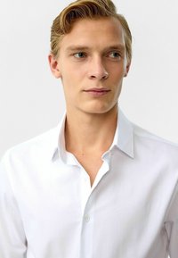 Witte overhemd met knopen en een klassieke kraag, gladde textuur en knooplijst; gedragen door een model met kort blond haar en een neutrale achtergrond.