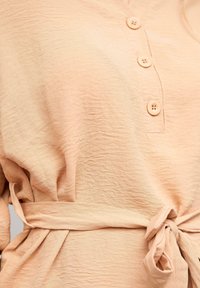 Peachfarvet bluse lavet af tekstureret stof, med knaplukning foran og en bundet talje. Blødt, let materiale med lette rynker.