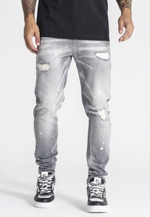 Mand iført lysegrå slidte jeans med revnede pletter og malingstænk, sorte og hvide sneakers og en sort skjorte.