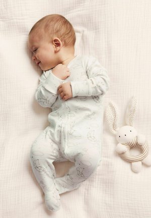 Un bébé dort sur une couverture à texture légère, vêtu d'une combinaison bleu pâle avec un motif de nuages et d'étoiles fantaisistes. Un doux jouet en forme de lapin rayé repose à proximité.