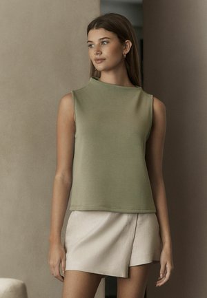 Top sin mangas de color verde oliva, confeccionado en tejido suave, con un escote alto y un corte relajado, combinado con una falda envolvente de un beige claro.