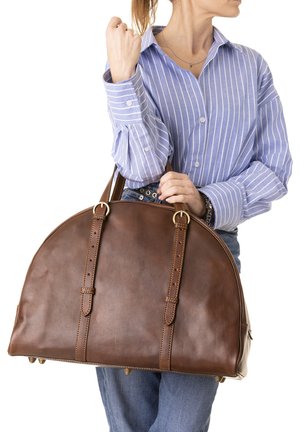 Donna con maglietta a righe che tiene una grande borsa di pelle marrone con doppi cinturini con fibbia e piedini in metallo sul fondo.