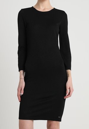 Kvinde iført en tætsiddende, langærmet, knælængde sort kjole med rund hals mod en ensfarvet lys baggrund.