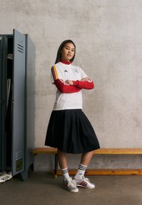 Λευκή κοντομάνικη μπλούζα με κόκκινες λεπτομέρειες και ρίγες, συνδυασμένη με μαύρη πλισέ φούστα. Γκρι αθλητικά παπούτσια με κόκκινες ρίγες και λευκές κάλτσες.