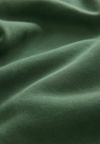 Tissu en coton vert avec une texture lisse, présentant des plis et des rides, mettant en avant sa douceur et sa qualité légèrement extensible.