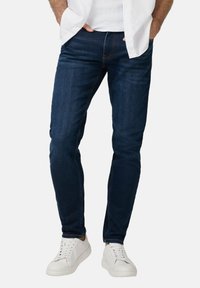 Granatowe dżinsy o slim fit, z pięcioma kieszeniami, kontrastowym przeszyciem, noszone z białymi trampkami. Gładka tekstura i delikatny połysk.