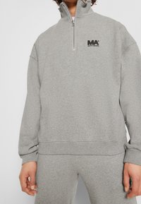 Sudadera gris con cremallera y cuello alto, puños y dobladillo acanalados. Presenta un logo negro en el pecho. Material de mezcla de algodón, ajuste relajado.