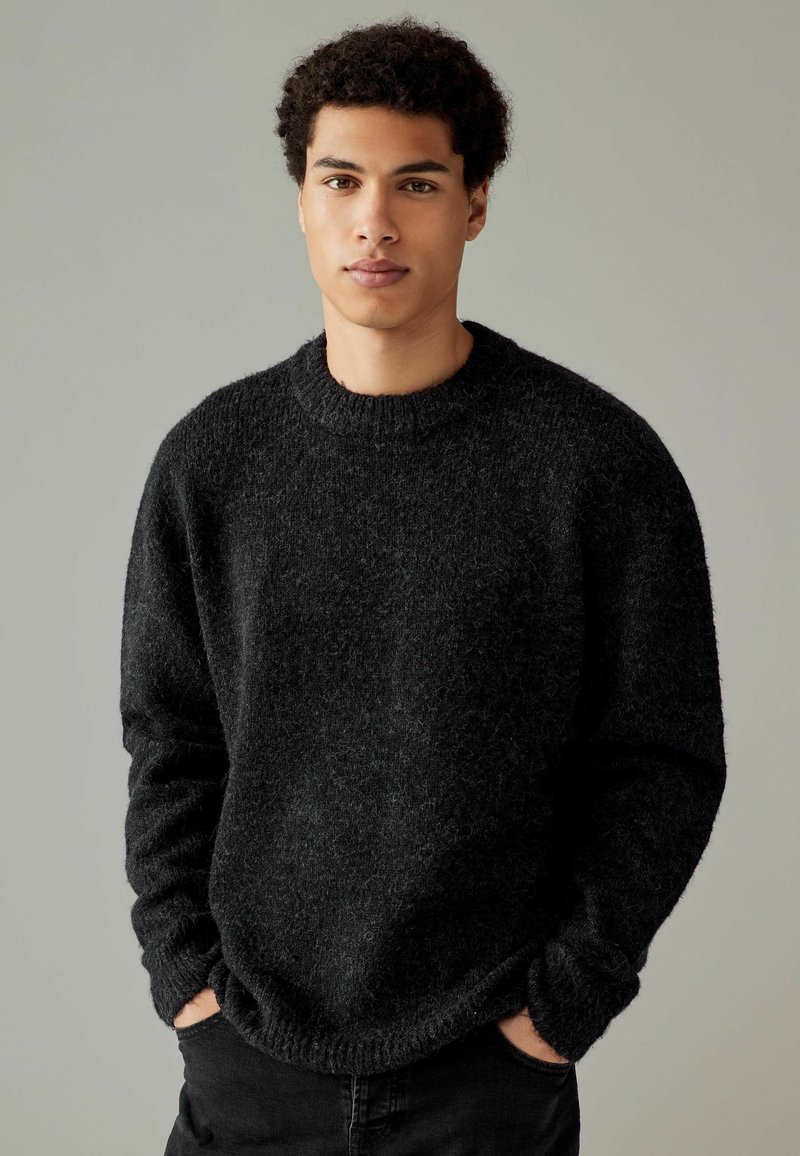 Schwarzer, strukturiert gestrickter Pullover mit Rundhalsausschnitt und elastischen Bündchen. Das Modell trägt ihn leger mit den Händen in den Taschen, vor einem neutralen Hintergrund.