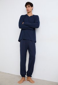 Marineblauw longsleeve shirt met een V-hals en een relaxed fit, gecombineerd met pyjamabroek met een patroon van een rode geometrisch ontwerp op een donkere achtergrond.