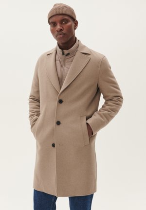 Beige wollen overcoat met grote revers en drie zwarte knopen, gedragen over een lichtgrijze pufferjack. Bevat zijzakken en heeft een soepele textuur.