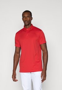 Polo vermelho feito de tecido suave, apresentando um colarinho, palas de botões e detalhes de design sutis nas mangas. Usado com calças brancas.