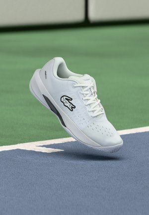 Chaussure de tennis blanche Lacoste avec logo noir, lacets blancs et semelle texturée flottant au-dessus d'un court de tennis bleu et vert.