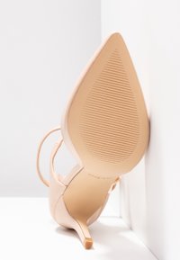 Chaussure à talon haut beige à bout pointu avec une fine bride, semelle orientée vers l'avant, positionnée contre un mur blanc d'angle.
