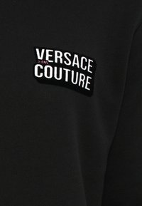 Sweater preto com um patch de logotipo bordado que diz "VERSACE COUTURE" com "JEANS" em rosa, criando um contraste texturizado.