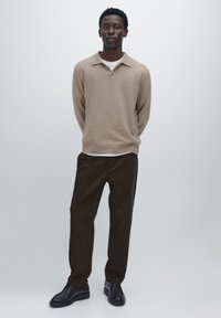 Man står med händerna bakom ryggen, iklädd en beige långärmad polo, vit undertröja, mörkbruna byxor och svarta skor mot en grå bakgrund.