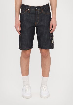 Shorts cargo in denim scuro con cuciture in contrasto, dettaglio delle tasche laterali e taglio dritto. Indossati con calzini bianchi e sneaker.