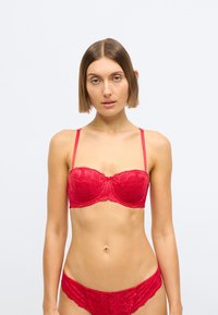 Červená čipkovaná podprsenka s kosticami a nastaviteľnými ramienkami. Má kvetinový vzor s vlnitými okrajmi a zodpovedajúce čipkované nohavičky.