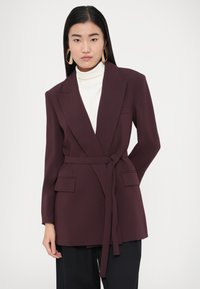 Bordó blézer övvel a deréknál, kerekített hajtókákkal és két elöl lévő zsebbel. Fehér garbó felett viselve, sötét nadrággal párosítva.