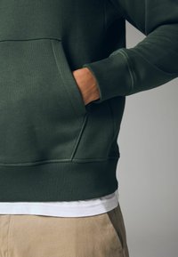 Sudadera verde oscura con un bolsillo tipo canguro; puños y dobladillo de canalé; usada sobre una camiseta blanca; el tejido parece suave con una textura lisa.