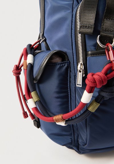 Sac à dos bleu avec poche avant, fermeture éclair argentée et cordons noués décoratifs rouges, marine, blancs, olive et marron attachés.