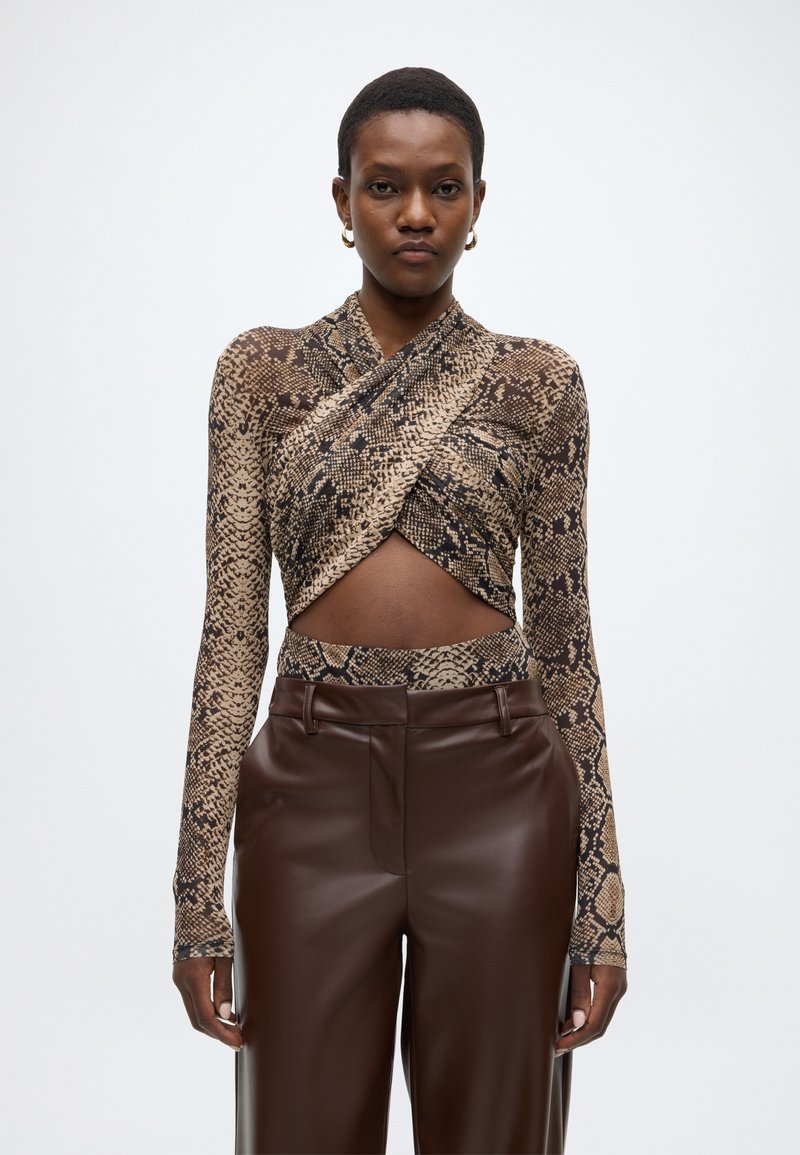 Langärmeliges, figurbetontes Oberteil mit Wickeldesign in Schlangenprint, ausgestattet mit einem tiefen V-Ausschnitt und einem Taillenausschnitt. Kombiniert mit braunen Lederhosen.